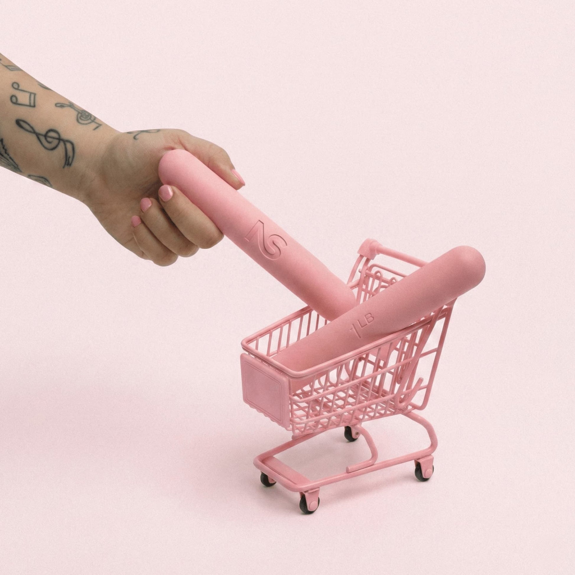 Main avec des tatouages plaçant des haltères roses Neo Sculpt Bars de 1 LB dans un mini chariot de supermarché rose.